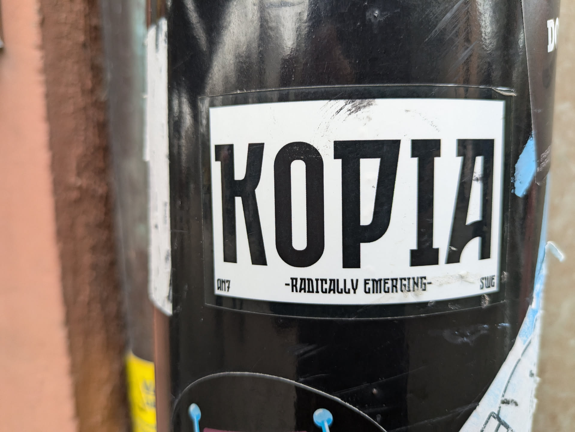 A black and white sticker with the word "KOPIA" in bold, uppercase letters. Below the word, it says "-Radically Emerging-". The sticker is attached to a black cylindrical object, possibly a pole or a pipe. 