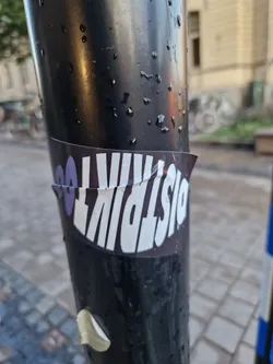 A sticker with the text DistriKt00 on a dark background. The sticker is applied to a dark-colored pole outdoors.
