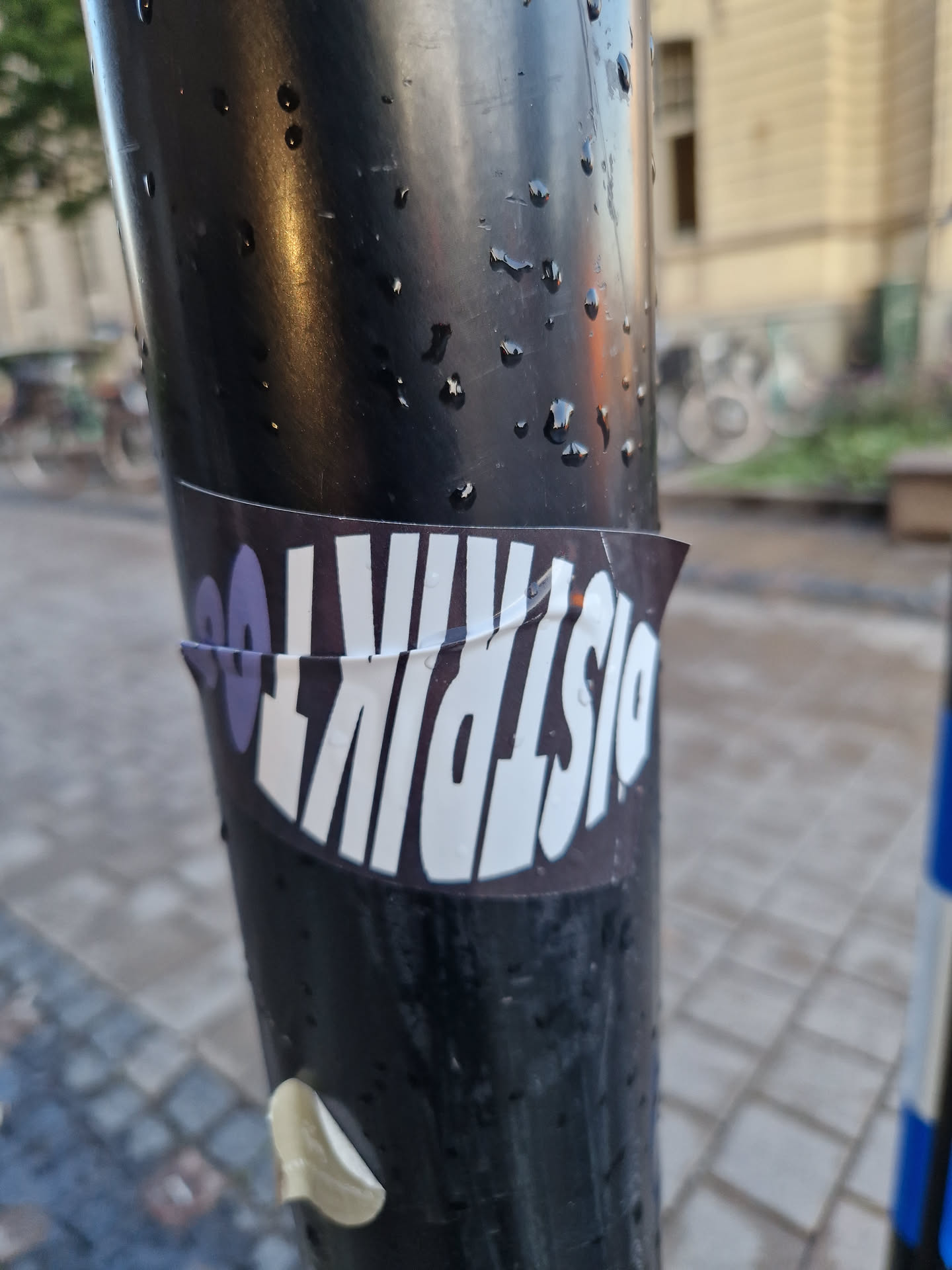 A sticker with the text DistriKt00 on a dark background. The sticker is applied to a dark-colored pole outdoors.