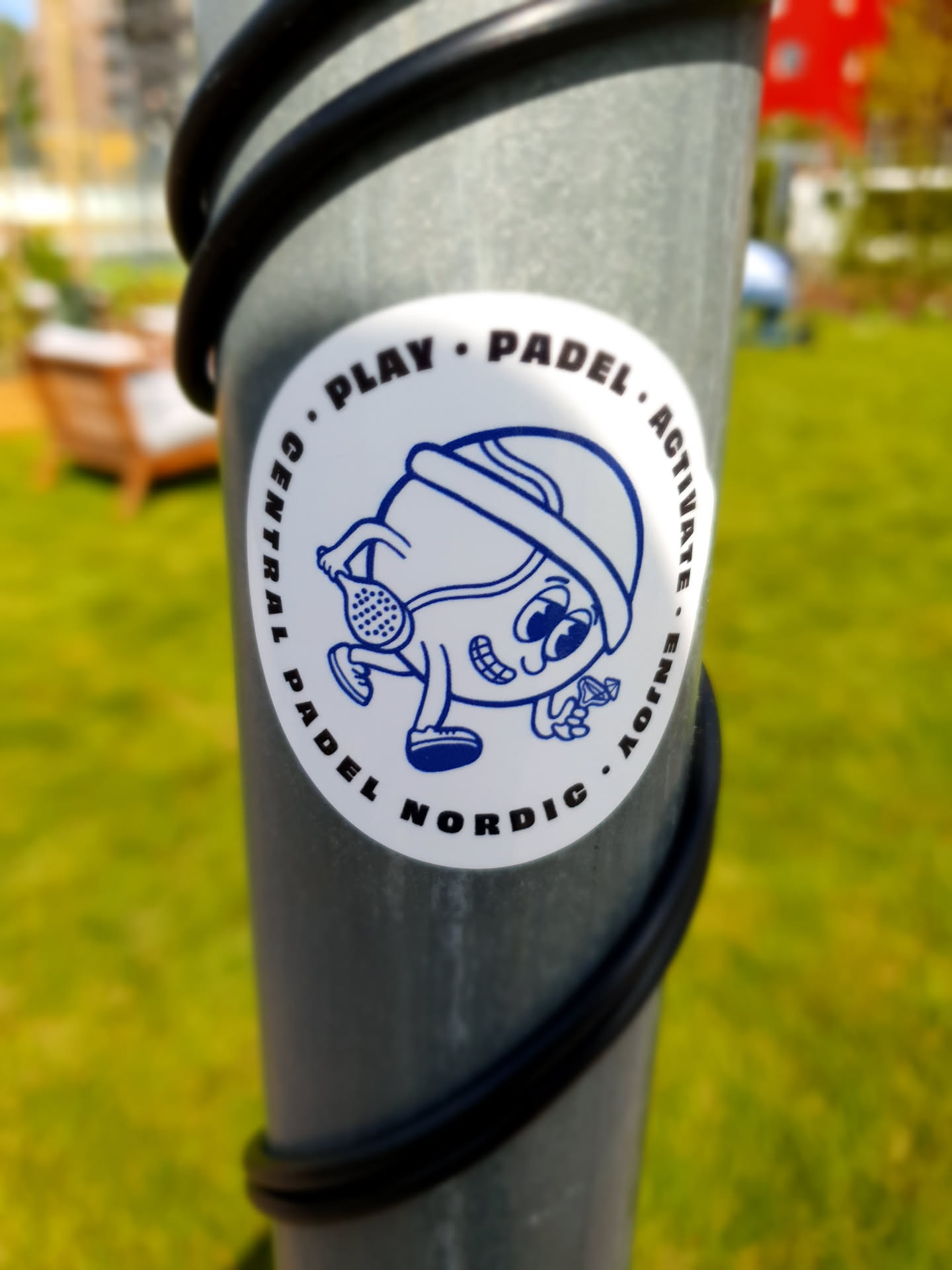 A round sticker with a cartoon character playing padel. The sticker is attached to a metal pole outdoors.