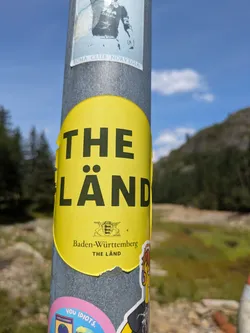 A vibrant yellow, oval-shaped sticker affixed to a grey metal pole. The prominent feature is the large, black, sans-serif text 'THE LÄND'. Below this, a smaller crest or coat of arms is visible, depicting two stylized deer or griffins supporting a shield. Beneath the crest, the words 'Baden-Württemberg' are printed in a smaller font, followed once more by 'THE LÄND'. The sticker shows some minor damage around its edges. The background is a blurred outdoor scene with trees, a valley, and a bright blue sky with some clouds.