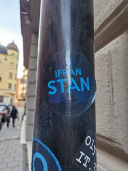 A round sticker with the text "IFRÅN STAN" in blue text on a dark background. The sticker is affixed to a dark-colored pole outdoors. The background shows a city street.