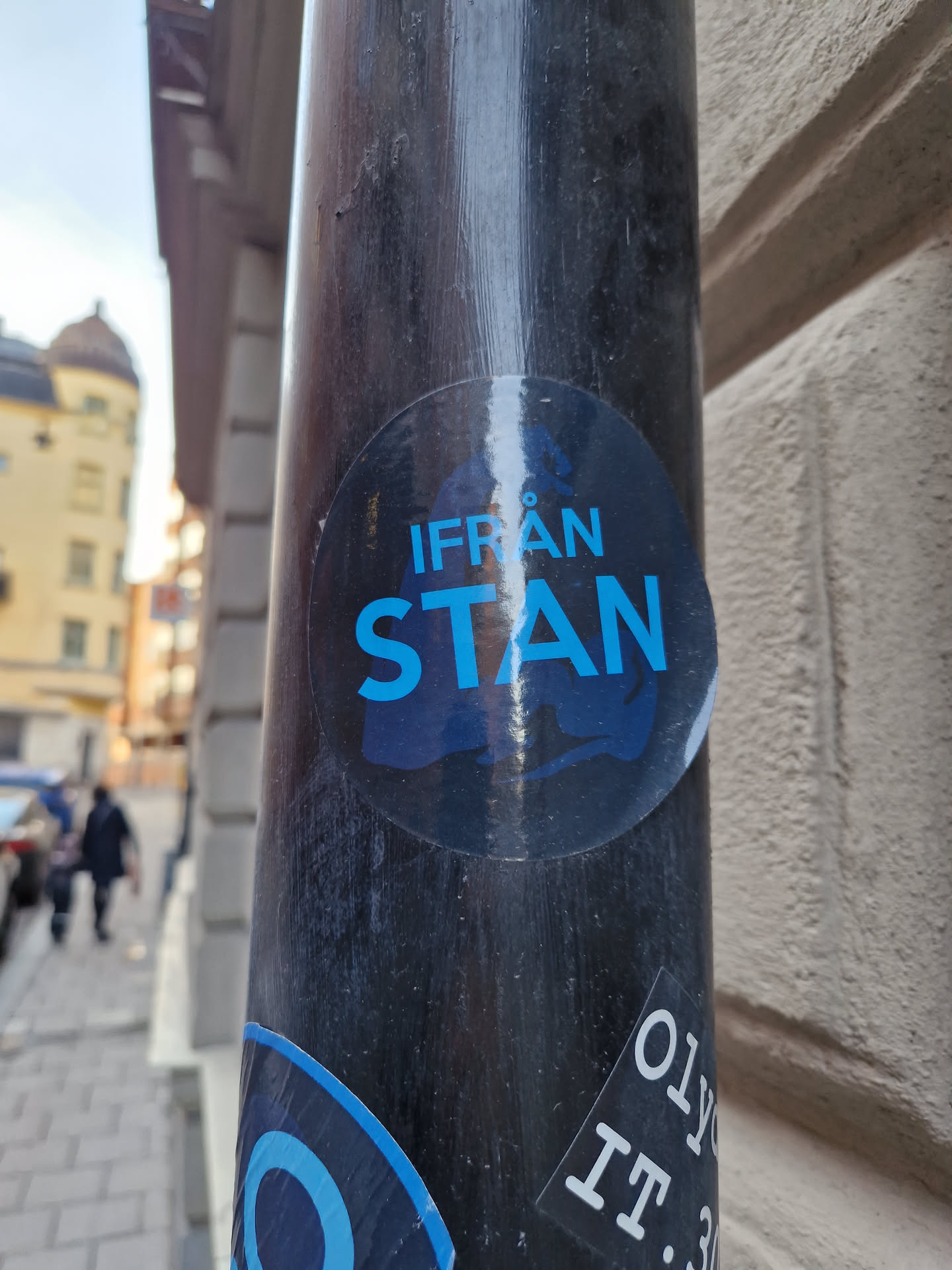 A round sticker with the text "IFRÅN STAN" in blue text on a dark background. The sticker is affixed to a dark-colored pole outdoors. The background shows a city street.