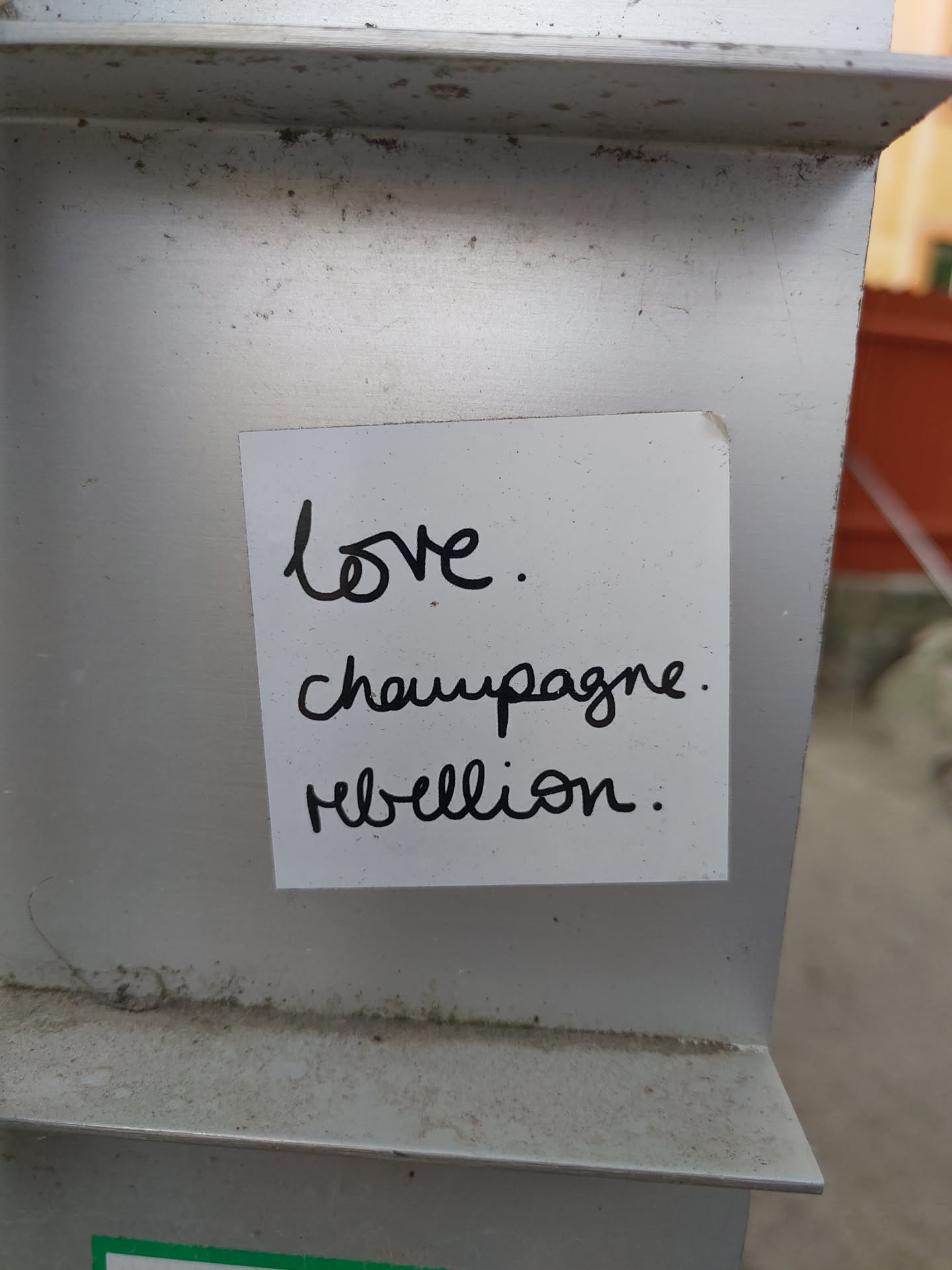 A small handwritten sticker affixed to a metallic surface. The sticker features the words 'Love. Champagne. Rebellion.' written in a cursive style.