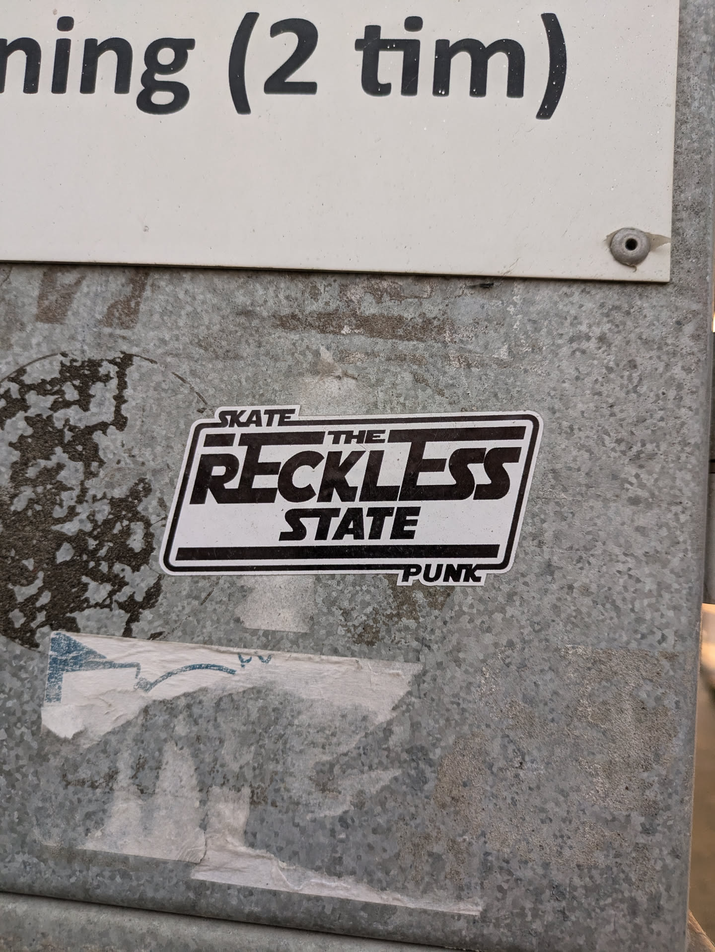 Black and white sticker with the text "Skate the Reckless State" in a stylized font. The word "Punk" is written below the main text. The sticker is attached to a metallic surface.