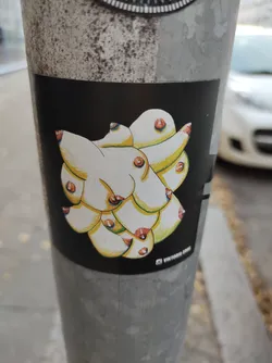 This sticker showcases a unique artistic interpretation of breasts. The style is graphic and slightly surreal, with the individual forms overlapping and interconnecting to create a cohesive abstract whole. The color palette is simple, relying on the contrast between the light yellow-beige of the breasts and the black background. The artwork’s overall effect is bold and eye-catching, despite its seemingly simple approach.  The artist's signature, "Vittoria G", adds a personal touch.