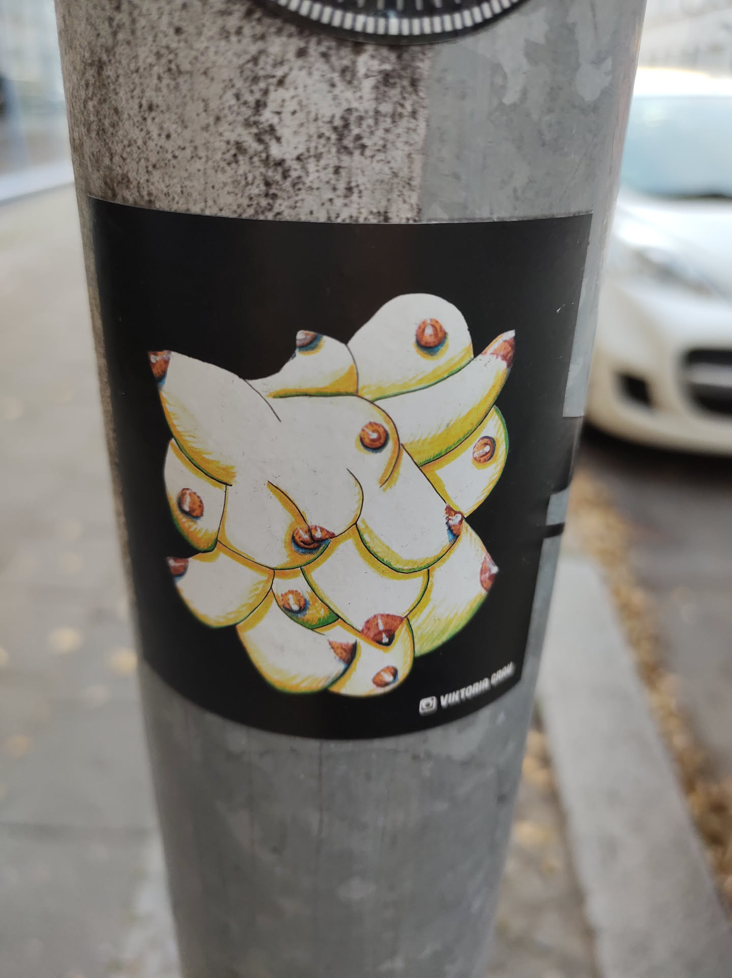 This sticker showcases a unique artistic interpretation of breasts. The style is graphic and slightly surreal, with the individual forms overlapping and interconnecting to create a cohesive abstract whole. The color palette is simple, relying on the contrast between the light yellow-beige of the breasts and the black background. The artwork’s overall effect is bold and eye-catching, despite its seemingly simple approach.  The artist's signature, "Vittoria G", adds a personal touch.