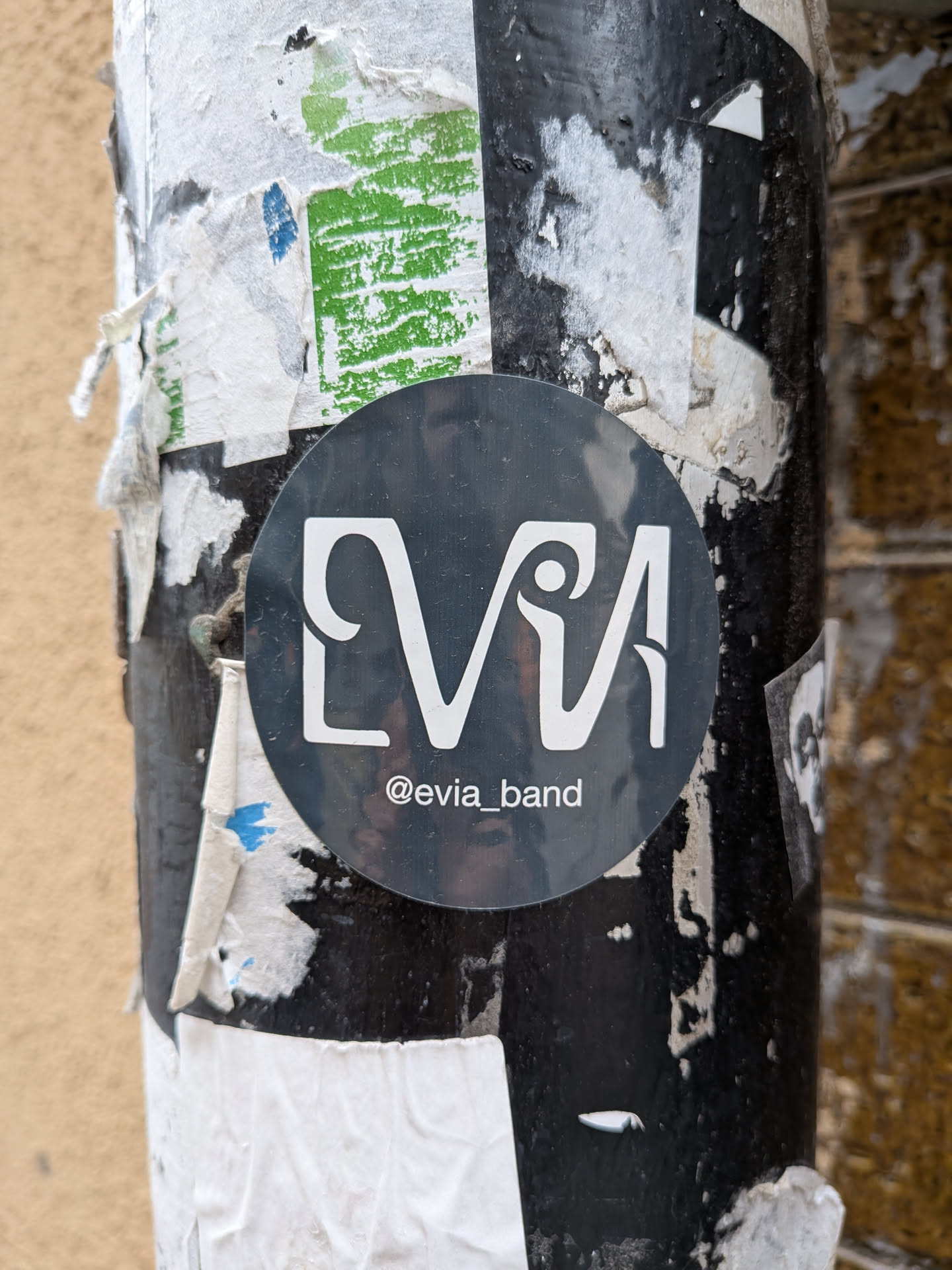 A circular sticker featuring a dark charcoal gray background. In the center, the word 'EVIA' is prominently displayed in a highly stylized, elegant white font. The 'E' and 'V' are connected, and the 'I' has a dot that resembles a head, while the 'A' has a unique, almost calligraphic flourish. Below the main logo, in smaller white text, is the social media handle '@evia_band'. The sticker has a slight sheen and is affixed to a heavily stickered and worn black pole.