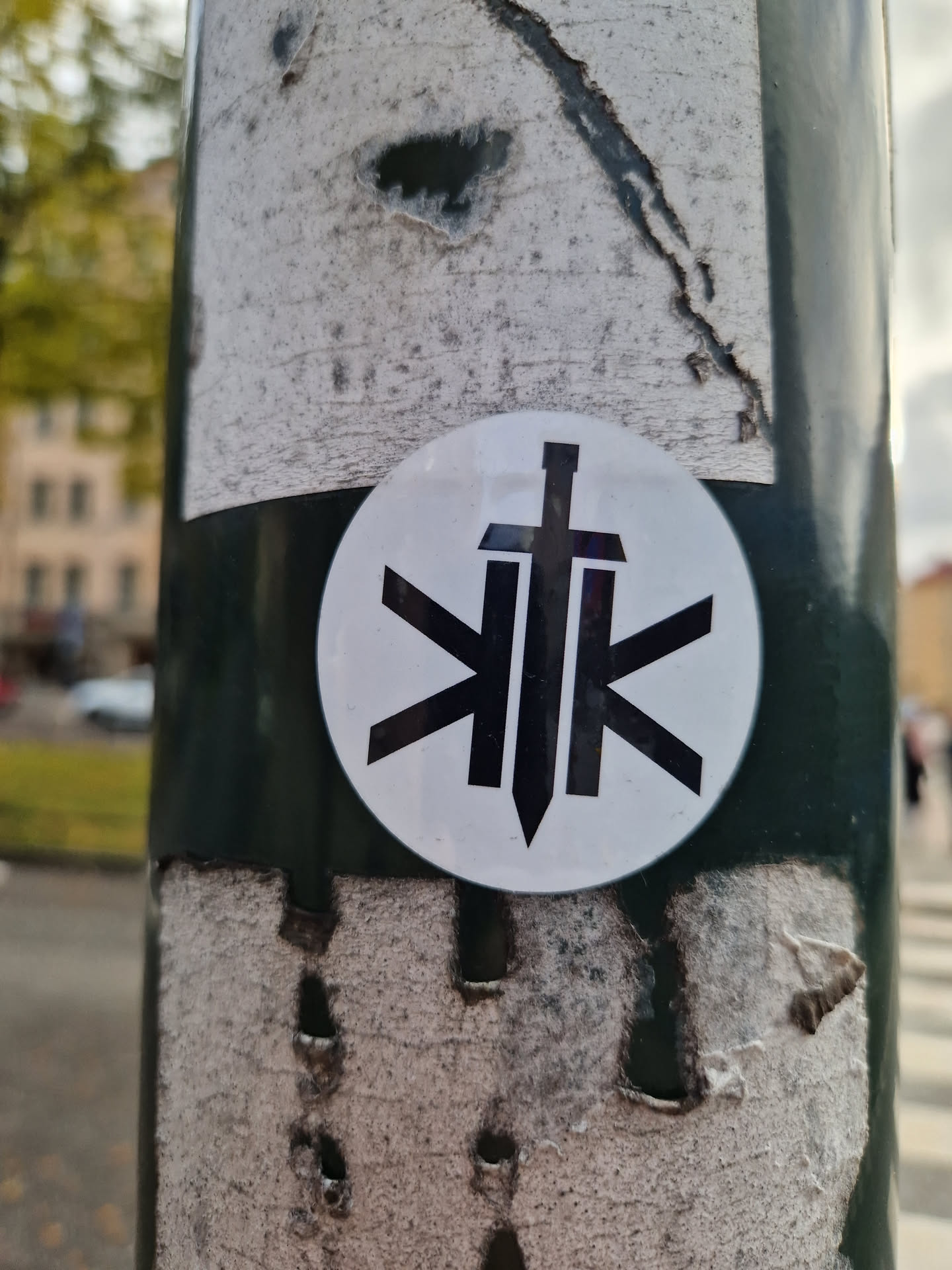 A round sticker with a black and white design. The central motif appears to be a stylized sword or dagger within a circular geometric pattern. The style is minimalist and graphic.