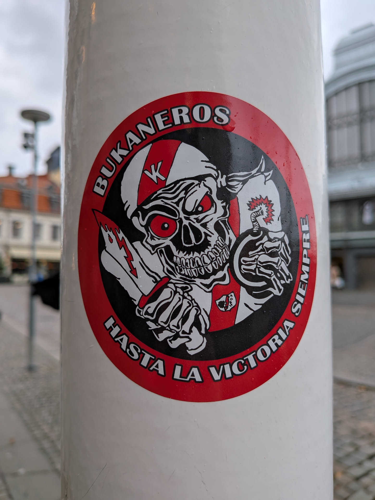 The sticker is circular with a red border. At its center is a detailed illustration of a skull wearing a helmet with a red and white striped pattern. The skull has glowing red eyes and a menacing grin. It is holding a bloody knife in its left hand and a lit bomb in its right hand. The background within the circle is black, with some white accents. Around the top of the circle, in white text with a black outline, are the words 'BUKANEROS'. Along the bottom of the circle, also in white text with a black outline, are the words 'HASTA LA VICTORIA SIEMPRE'. There is a small crest-like emblem on the skull's chest.