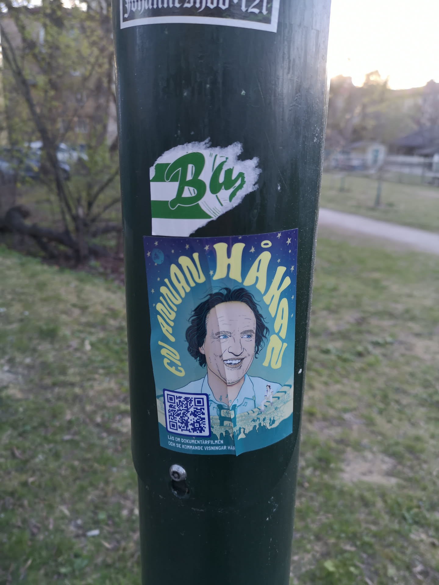 A vibrant, rectangular sticker featuring a stylized, smiling portrait of a man with dark, curly hair and blue eyes, likely Håkan Hellström. The portrait is split, with one side more detailed and the other slightly simpler. Above his head, the text 'EN ANNAN HÅKAN' is written in curved yellow letters against a dark blue, star-filled background. Below the portrait, a light blue section depicts a large crowd of silhouetted figures, suggesting a concert or event. A prominent blue and white QR code is located in the bottom left, with small black text beneath it on a light blue strip, reading 'LÄS OM DOKUMENTÄRFILMEN OCH SE KOMMANDE VISNINGAR HÄR'. The sticker has a slightly worn appearance, consistent with being affixed to a pole.
