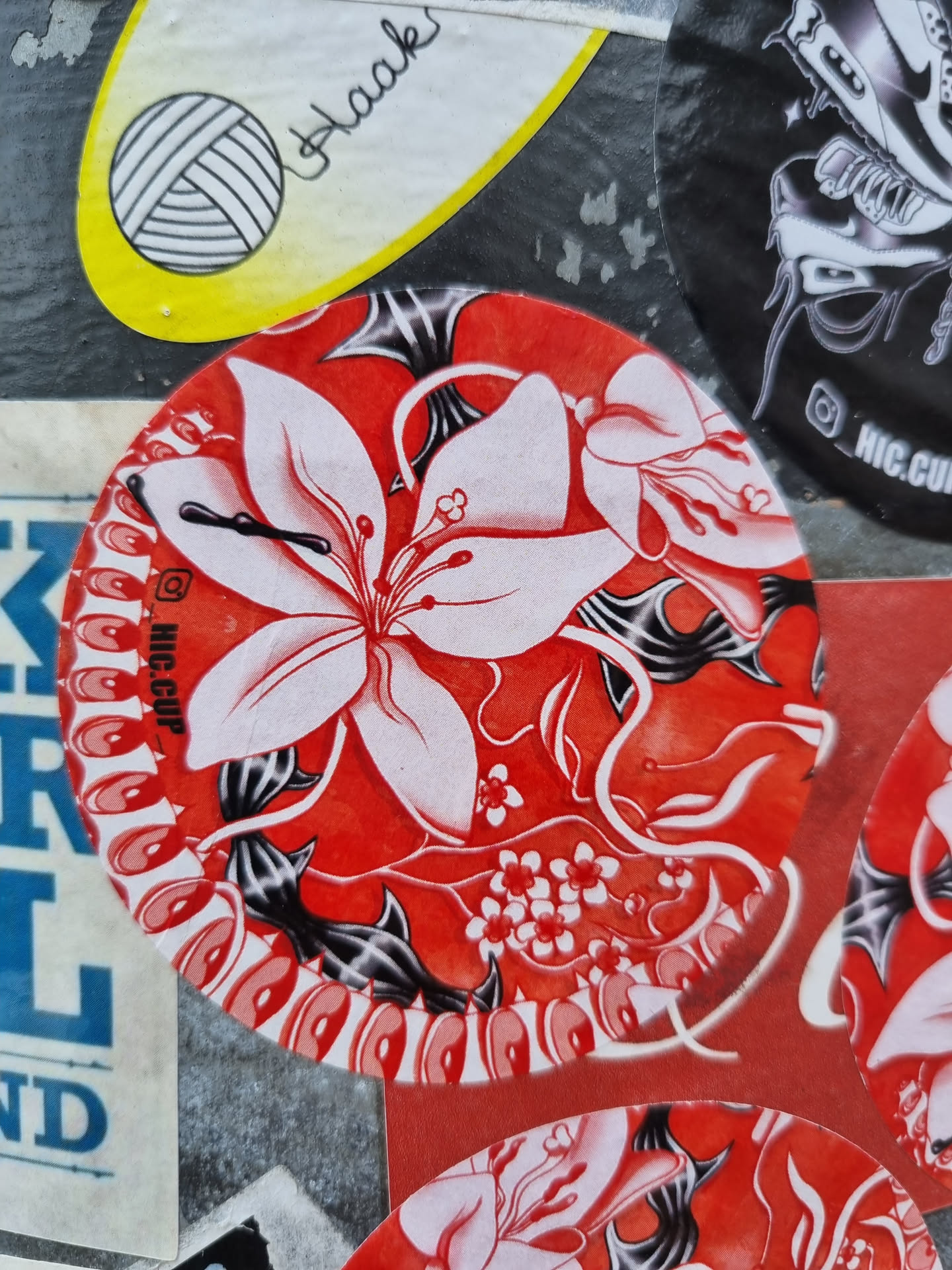 Round sticker featuring a stylized floral pattern in red, white, and black. The main design element is a large white lily-like flower with intricate details. The background is a bold red with black swirling lines and smaller white flowers.  There is a small Instagram logo visible on the sticker.