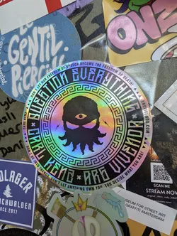 Holographic iridescent sticker with a circular design featuring a stylized figure with an eye and tentacles, surrounded by a Greek key pattern and text.  The text reads, "Question Everything," "Carl Kenzie Are Vivendo," and a statement about freedom of speech. A QR code is present, linking to a livestream and social media.