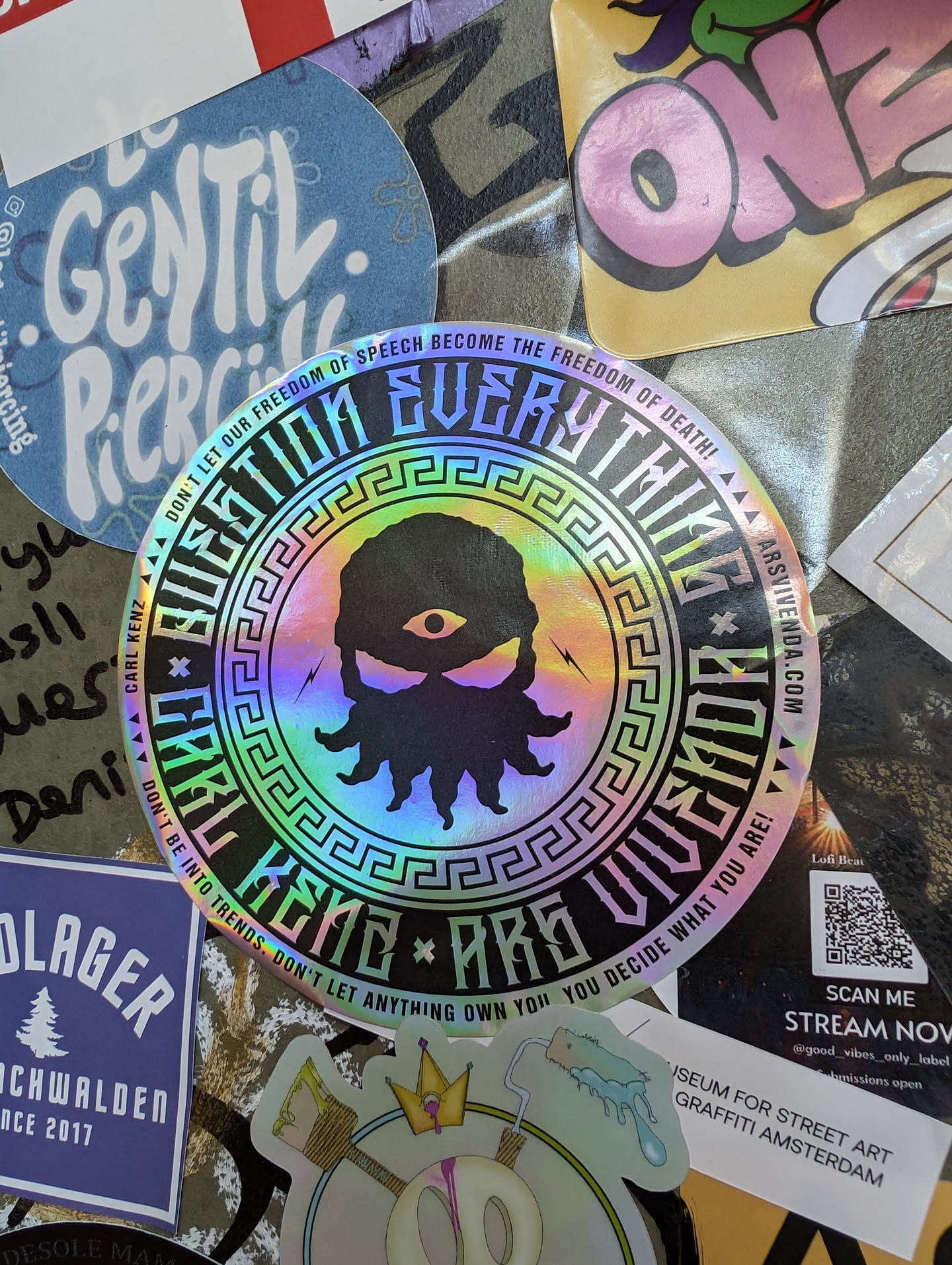 Holographic iridescent sticker with a circular design featuring a stylized figure with an eye and tentacles, surrounded by a Greek key pattern and text.  The text reads, "Question Everything," "Carl Kenzie Are Vivendo," and a statement about freedom of speech. A QR code is present, linking to a livestream and social media.