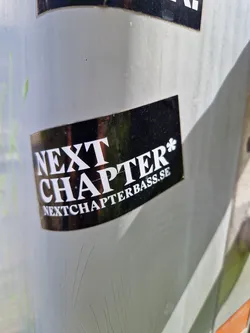 A black and white sticker with the text "NEXT CHAPTER*" and the website address "NEXTCHAPTERBASS.SE" below it. The sticker has a glossy finish.