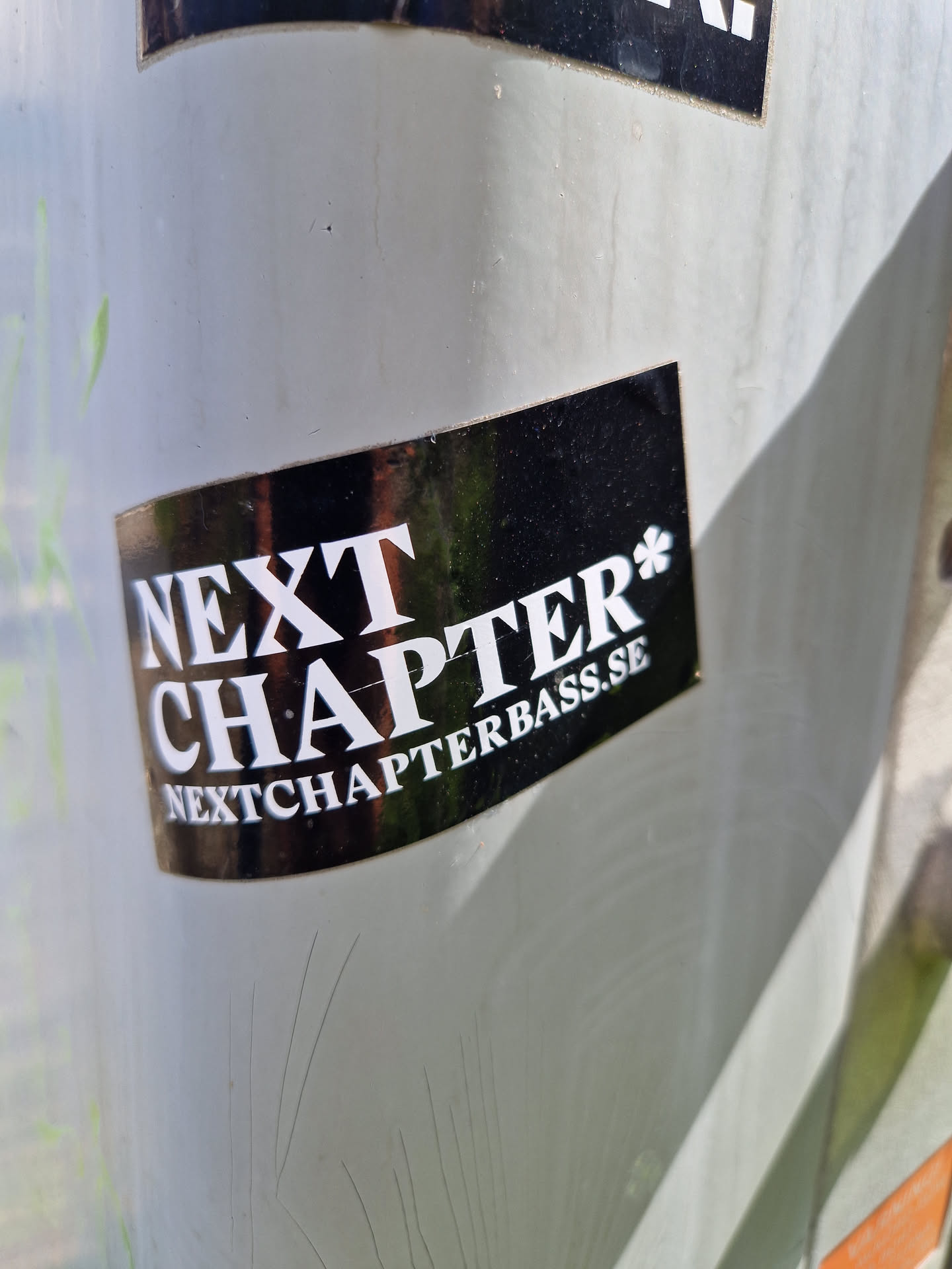 A black and white sticker with the text "NEXT CHAPTER*" and the website address "NEXTCHAPTERBASS.SE" below it. The sticker has a glossy finish.
