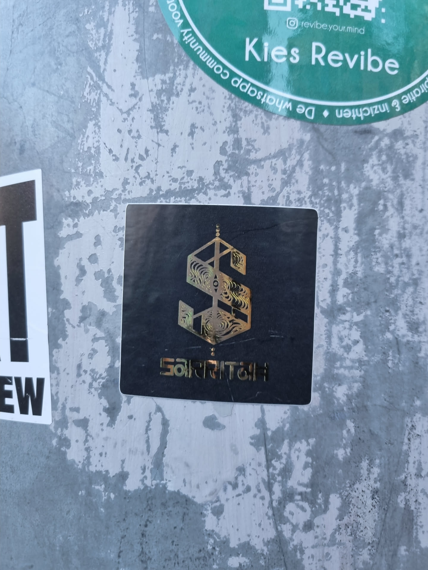 A black square sticker with a gold geometric design and the word Sakritai underneath.  The design features a stylized 'S' with intricate details. 
