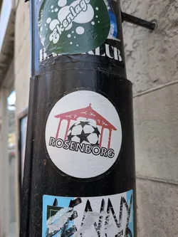 A circular white sticker featuring a stylized red pavilion or gazebo-like structure. Beneath this structure, a black and white soccer ball is depicted, resembling the UEFA Champions League ball with its characteristic star pattern. The word "ROSENBORG" is written in bold, black, sans-serif capital letters, curving along the bottom edge of the circle, partially overlapping the soccer ball.