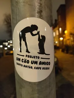 Round white sticker with black silhouettes of a person and a dog high-fiving each other. The text below indicates it's a project called "Um Cão Um Amigo" from Santo Antão, Cape Verde.