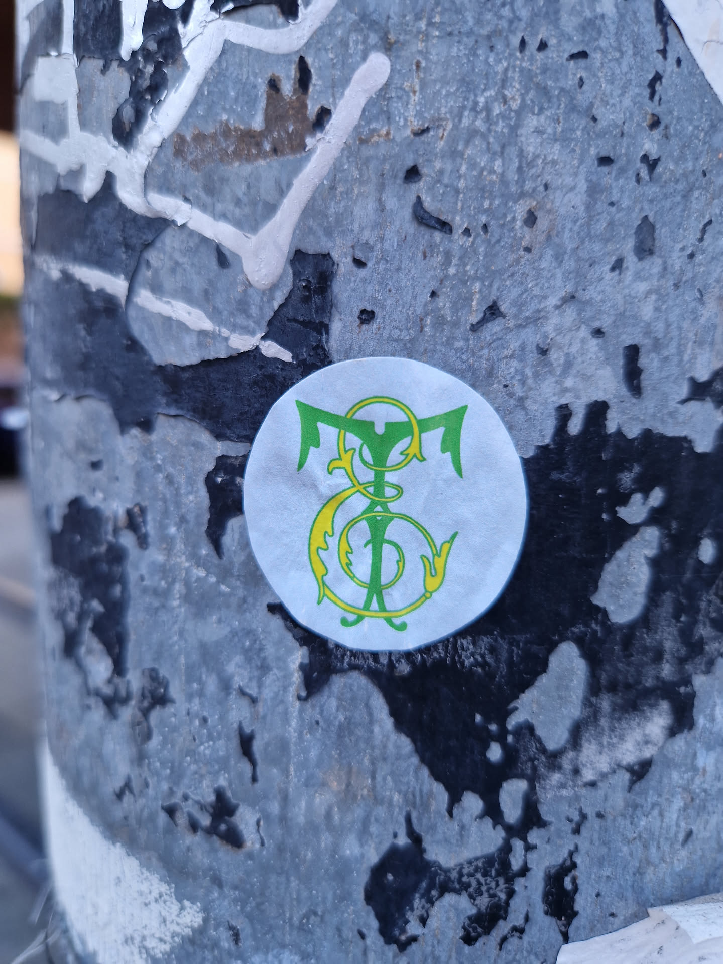 A round sticker with a monogram featuring intertwined letters or symbols in green and yellow on a white background.  The style is reminiscent of vintage or heraldic designs. It's affixed to a weathered surface.