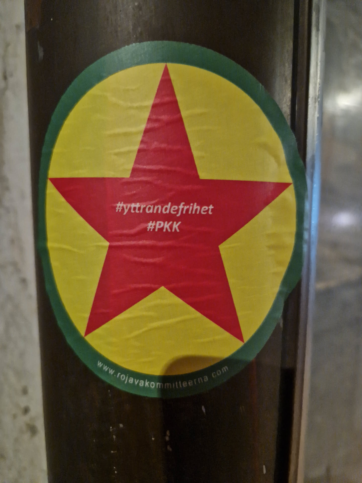 A round sticker with a red star in the center on a yellow background. The text #yttrandefrihet #PKK is visible on the star. At the bottom, a website address is printed: www.rojavakommitterna.com