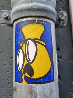 A sticker featuring a cartoon-like yellow character wearing large glasses and a hat. The style is simple, with bold outlines and flat colors. The character is set against a bright blue background, making it stand out. 
