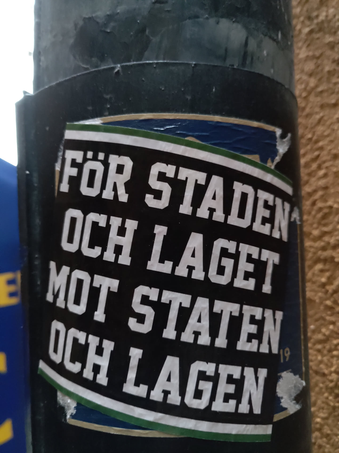 A black and white sticker with green and gold accents. The text is in Swedish and translates to "For the city and the team against the state and the teams". The number 19 is visible in the lower right corner. The sticker is affixed to a dark gray pole.