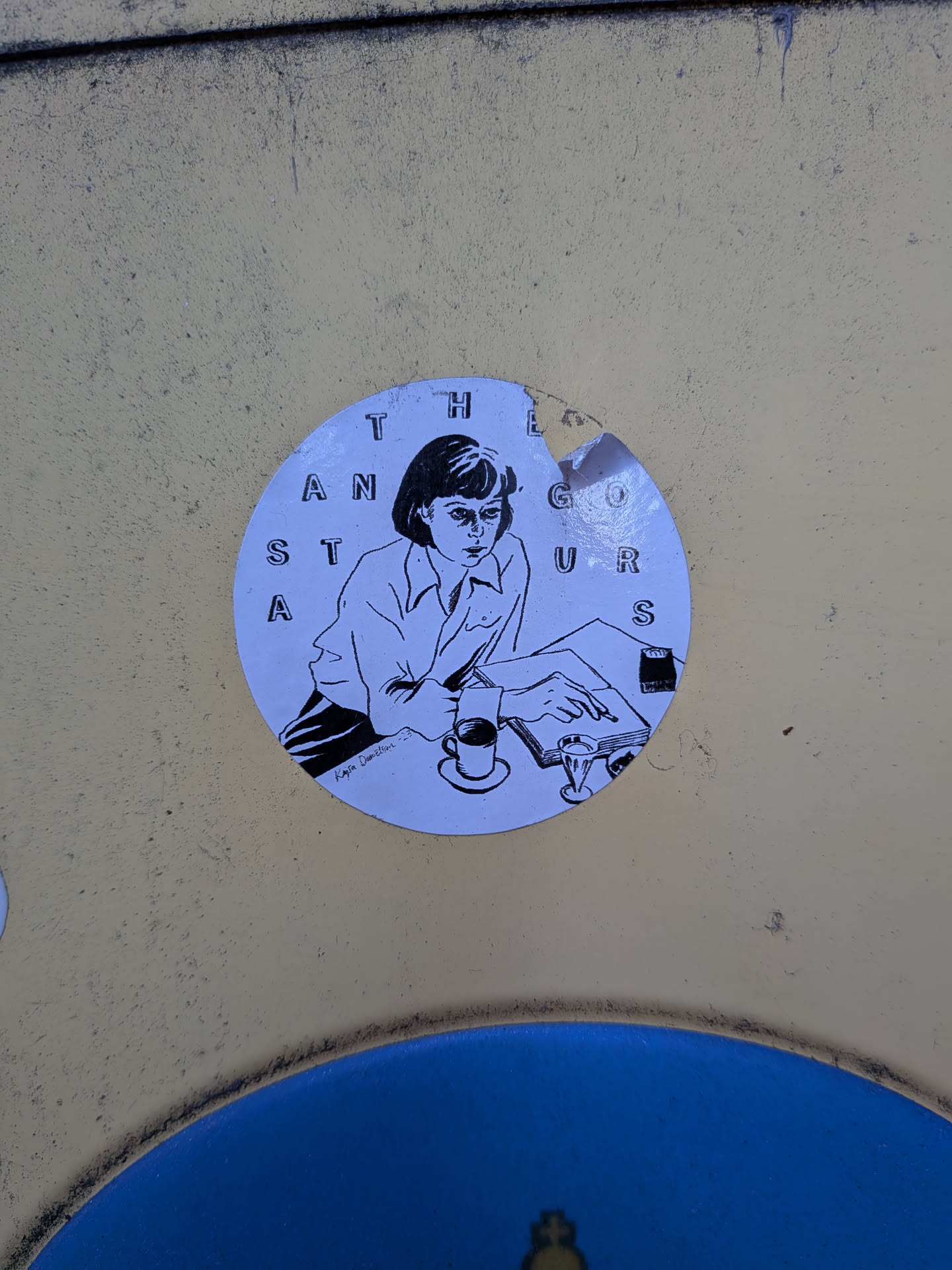 The sticker is circular and features a black and white illustration of a woman sitting at a desk, looking intently at a book. She is wearing a collared shirt and has short, dark hair. To her left, there is a coffee cup and saucer, and to her right, a small glass. The background of the sticker is a light blue. Around the edge of the circle, the letters 'T H E A N S T A G O U R S' are arranged in a circular pattern. There is a small signature at the bottom left of the illustration that appears to read 'Kajsa Damelien'. The sticker shows some signs of wear and tear, with a small piece missing from the top edge.