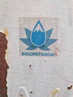 A sticker with a logo depicting a blue water droplet containing a mountain peak, set within a stylized flower-like design. The text 'DOLOMITIGROW' is present below the logo.