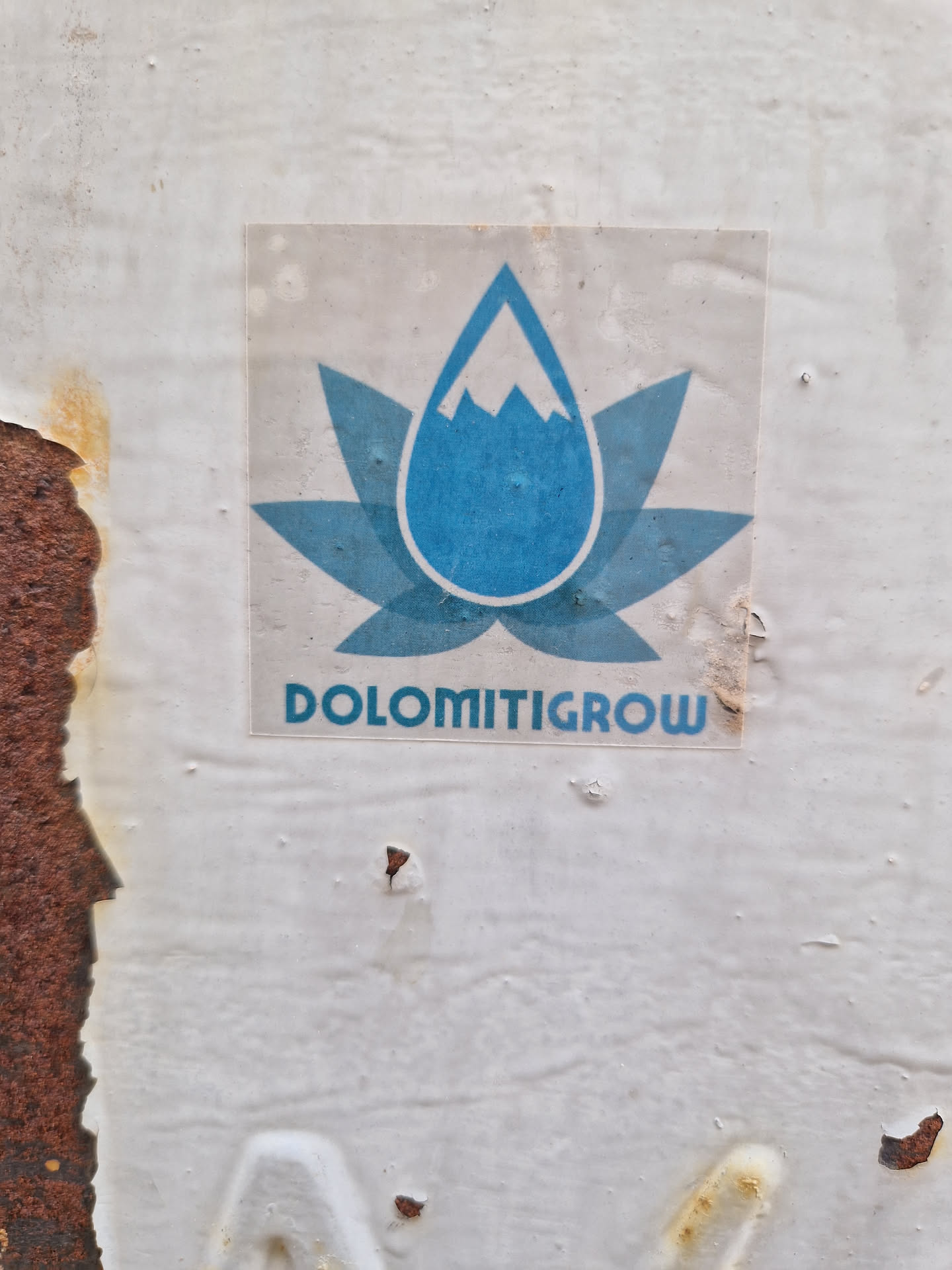 A sticker with a logo depicting a blue water droplet containing a mountain peak, set within a stylized flower-like design. The text 'DOLOMITIGROW' is present below the logo.