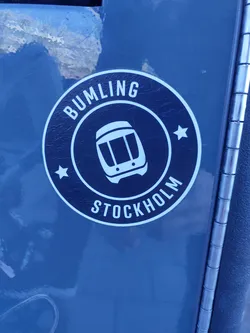 A round black and white sticker with the text "BUMLING" and "STOCKHOLM" in a circle, with a small train icon in the center and two stars on each side. The sticker is applied to a dark gray surface.