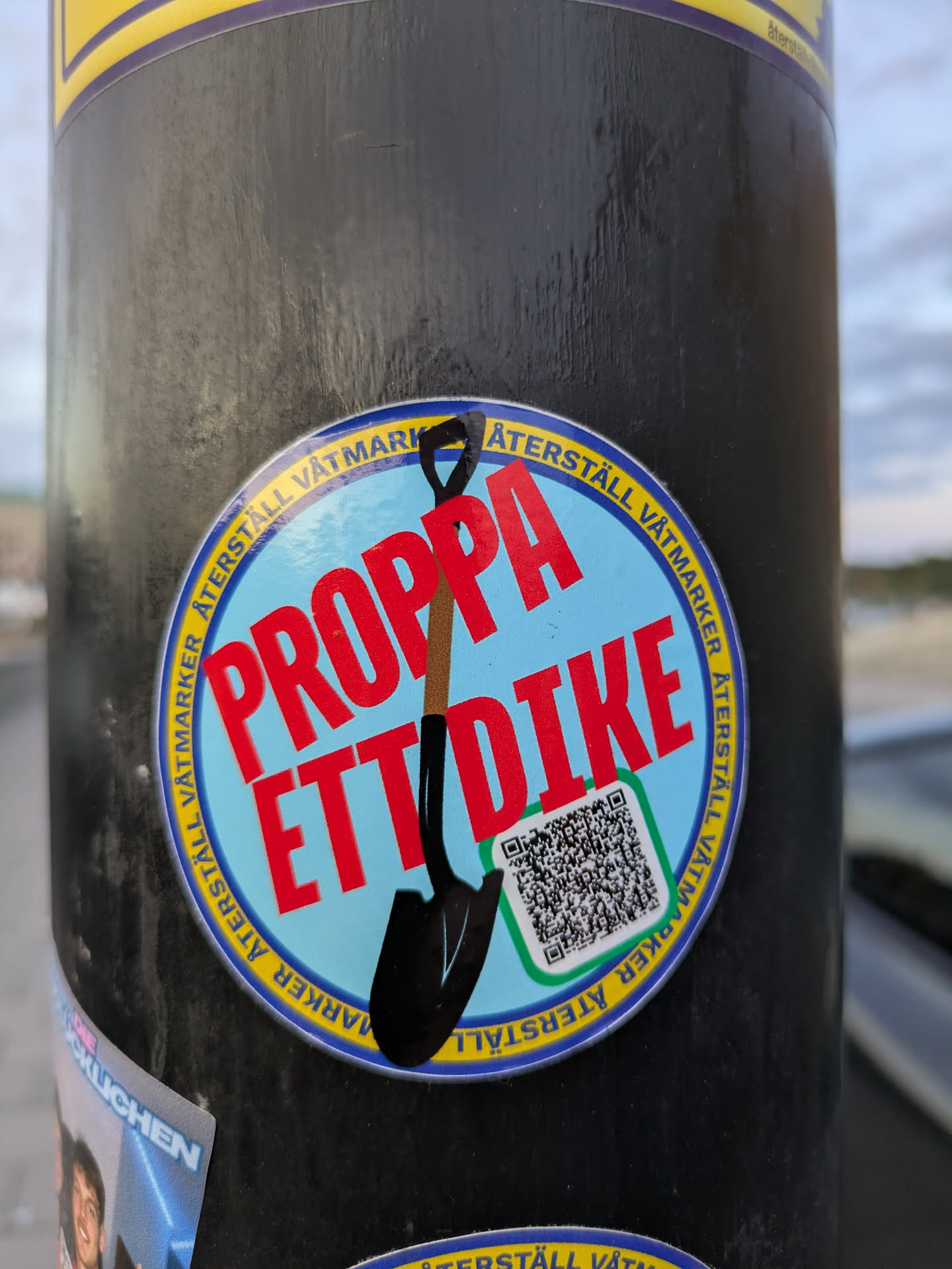 The central sticker is circular with a light blue background. It features a stylized black shovel with a brown handle pointing diagonally downwards. Bold, red text across the sticker reads 'PROPPA ETT DIKE'. Around the circumference of the sticker, in yellow text on a blue band, are the words 'ÅTERSTÄLL VÅTMARKER' repeated. A green-bordered square containing a QR code is positioned in the lower right quadrant of the sticker.