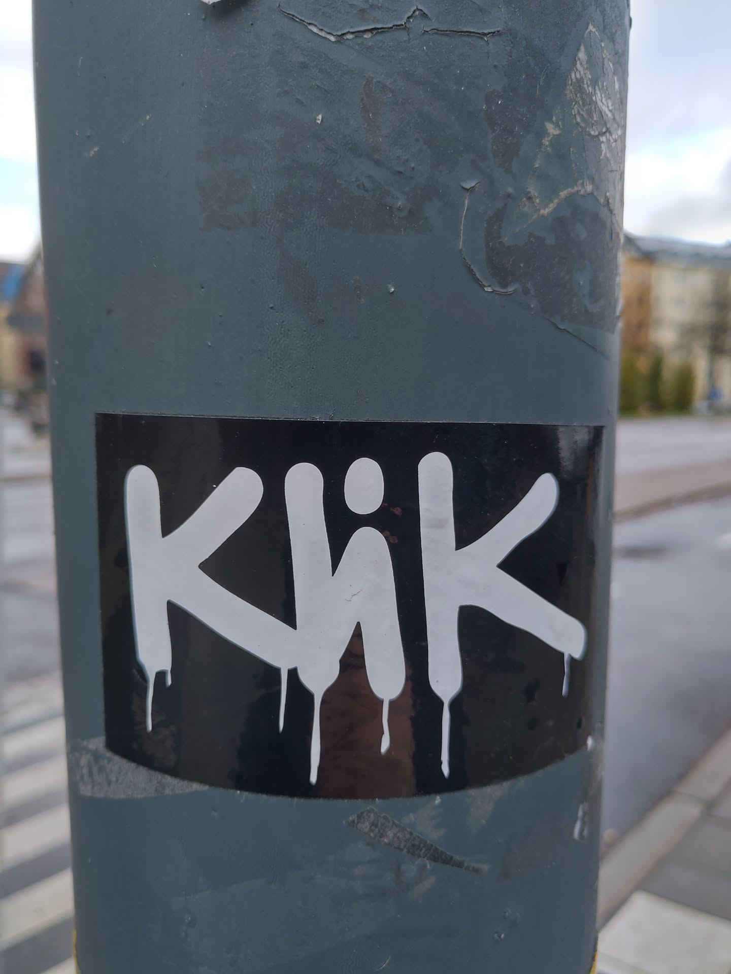 Black and white sticker with the word Khik written in a graffiti style with dripping paint effect. The sticker is placed on a grey pole.