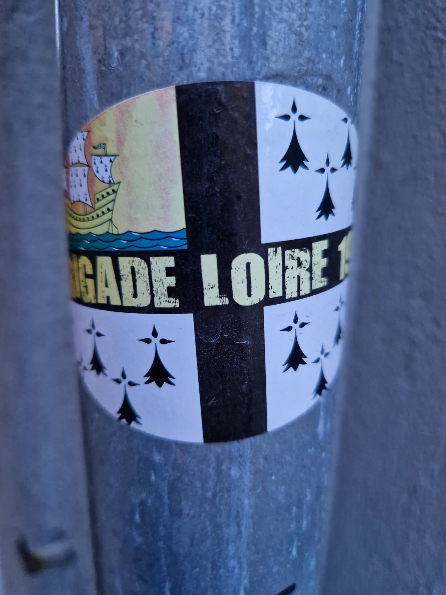 Round sticker with a design that looks like a heraldic emblem. It's divided into quadrants, with a ship on one side, ermine spots on two others, and a central black section with partially visible text. The visible text reads 'GADE LOIRE 1'.