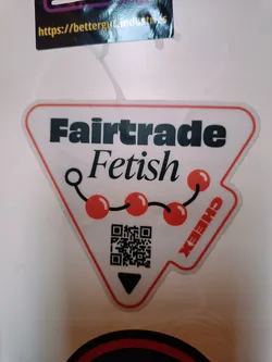 White triangle sticker with black text that says Fairtrade Fetish. It also contains a chain of red cherries and a QR code at the bottom. There is also a small black triangle pointing down. The sticker is slightly worn and has some creases.