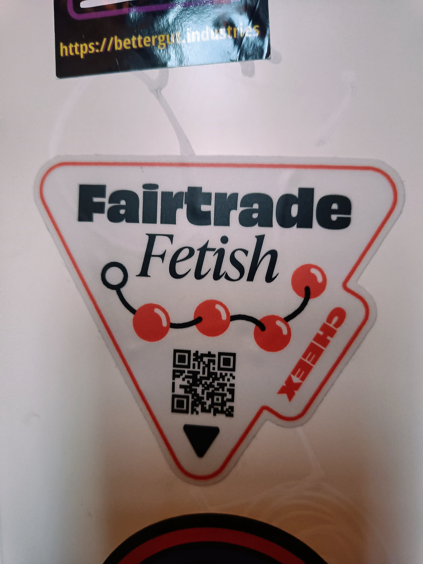 White triangle sticker with black text that says Fairtrade Fetish. It also contains a chain of red cherries and a QR code at the bottom. There is also a small black triangle pointing down. The sticker is slightly worn and has some creases.
