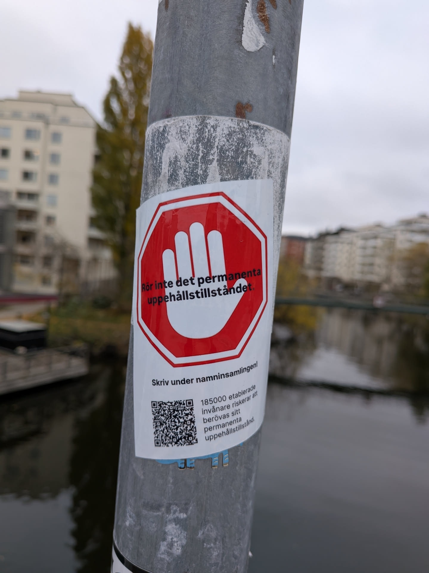 The sticker is predominantly white with a prominent red octagonal border, resembling a stop sign. Inside the octagon, a white open hand with fingers spread is depicted. Below the stop sign graphic, there is text in Swedish and a QR code. The text reads 'Rör inte det permanenta uppehållstillståndet.' (Do not touch the permanent residence permit.) and 'Skriv under namninsamlingen!' (Sign the petition!). Below this, there is smaller text and the QR code. The QR code is black and white, and appears to be scannable. The sticker is affixed to a metal pole.