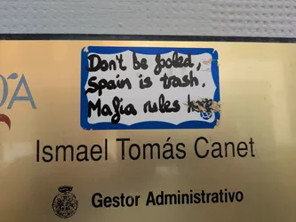 A sticker with a blue border and handwritten text in black marker. The sticker reads, "Don't be fooled, Spain is trash. Mafia rules." It's placed on a sign that reads "Ismael Tomás Canet, Gestor Administrativo."