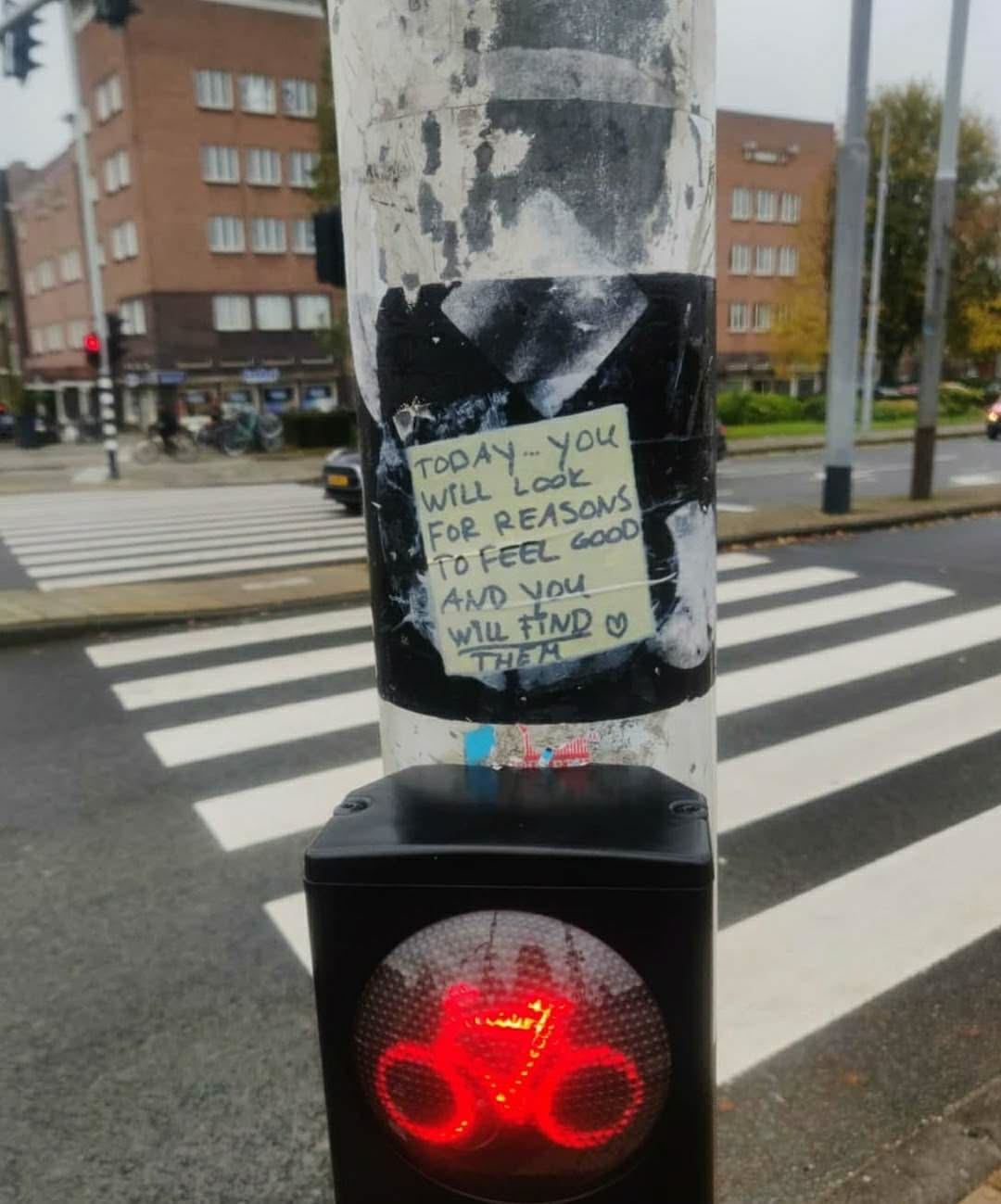 A yellow sticky note with a positive affirmation stuck on a pole at a crosswalk. The text on the note reads:
"TODAY... YOU
WILL LOOK
FOR REASONS
TO FEEL GOOD
AND YOU
WILL FIND
THEM"