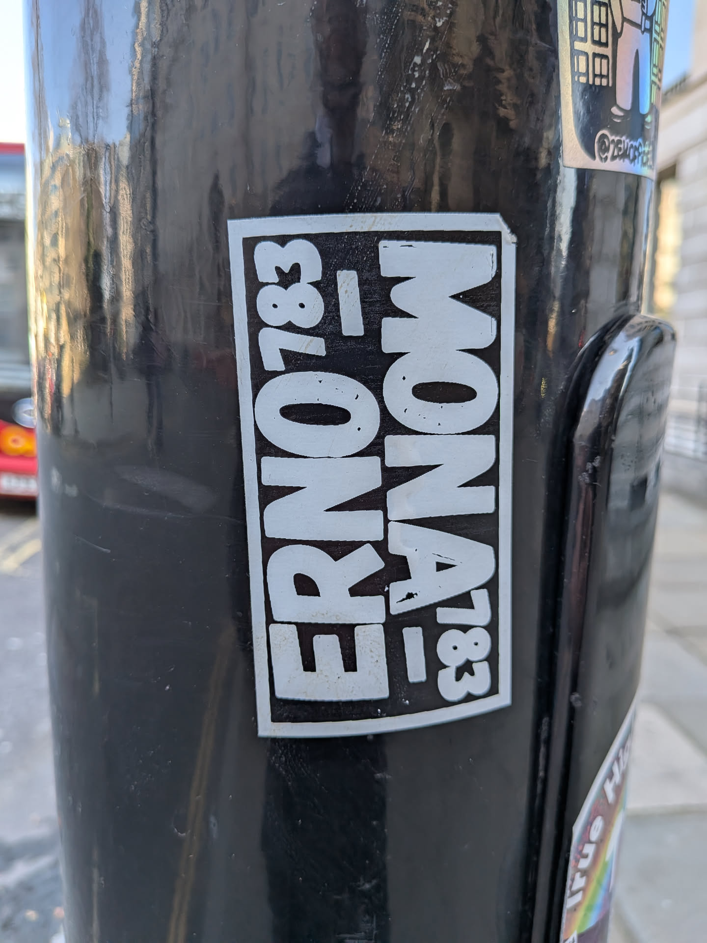 A rectangular, black and white sticker adhered to a dark, reflective pole. The sticker features bold, sans-serif white text on a solid black background, enclosed by a thin white border. The text is arranged in two vertical columns. The left column clearly reads 'ERNO' stacked vertically, followed by '783' stacked vertically. The right column mirrors this structure but is oriented upside down relative to the left column, displaying 'MONA' stacked vertically (with the 'A' at the top and 'M' at the bottom) and '783' stacked vertically (with the '3' at the top and '7' at the bottom). The overall design is clean, graphic, and industrial in feel, with some minor wear and tear visible on the white elements, suggesting it's a physical sticker.