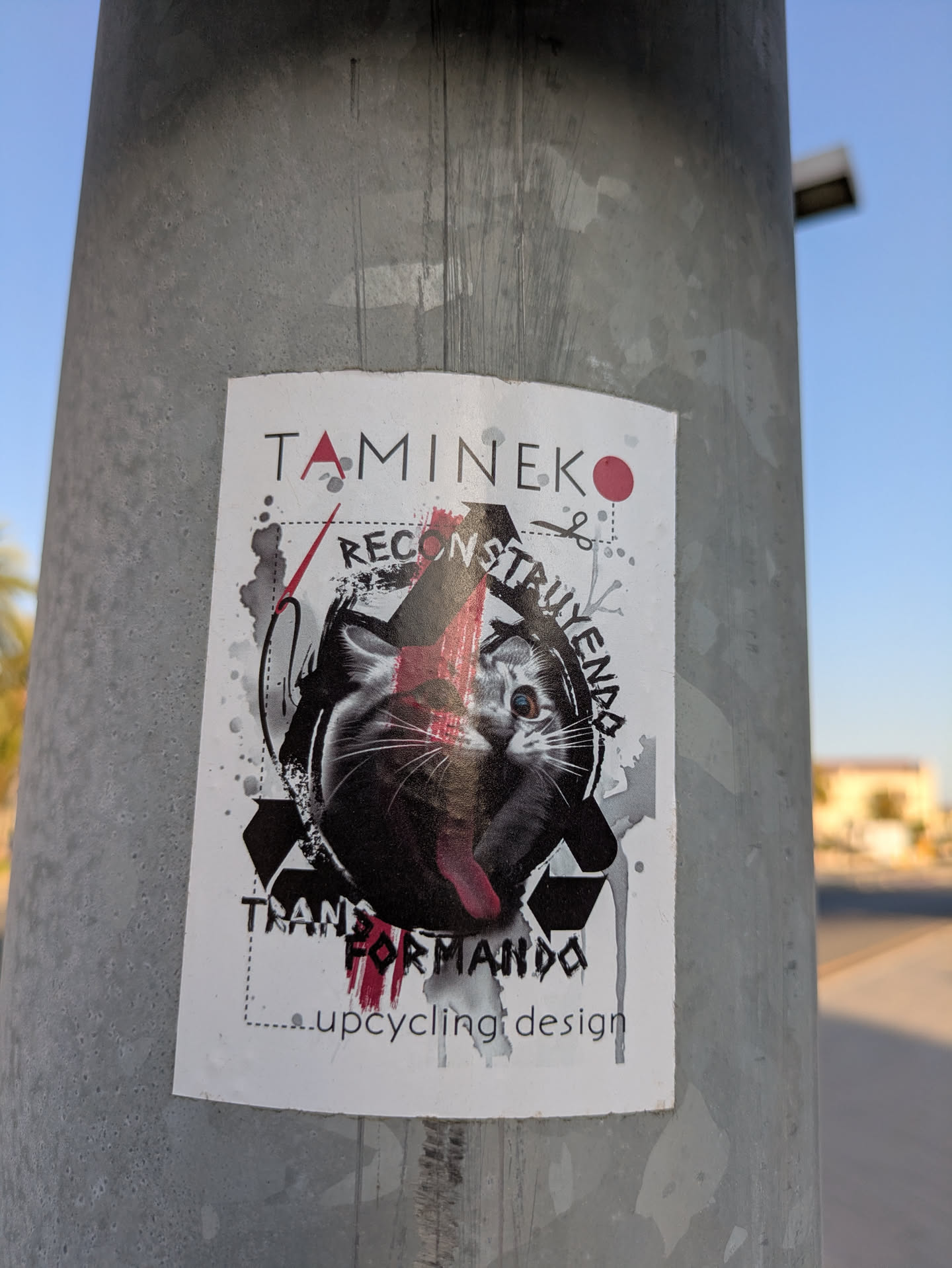 A white sticker on a metal pole features an image of a cat and the text 'Tamineko Reconstruyendo Transformando upcycling design'. The art is done with black and red paint.