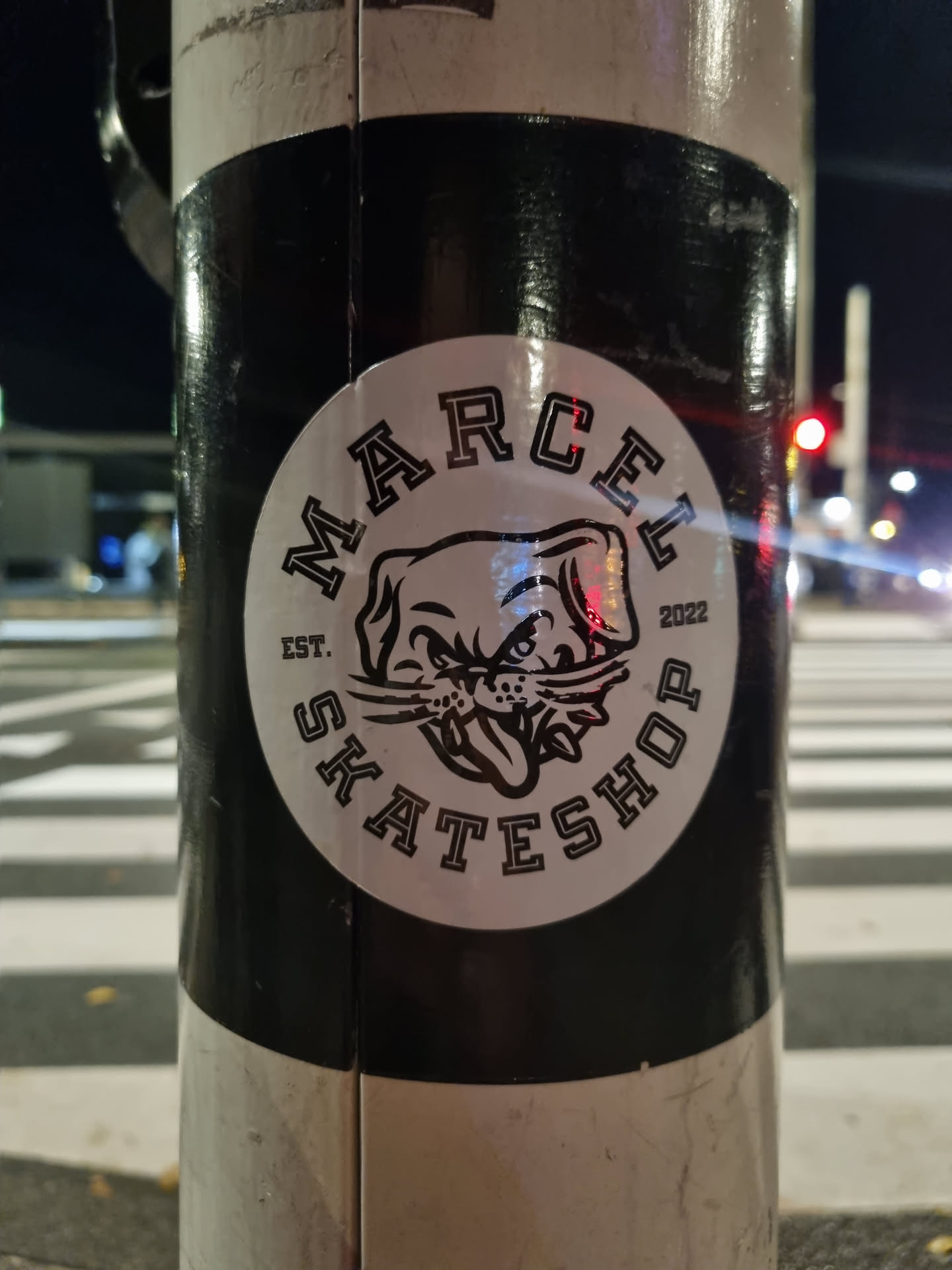 A black and white circular sticker featuring a dog mascot with the text 'Marcel Skateshop' and 'Est. 2022'. The sticker is affixed to a pole outdoors at night.