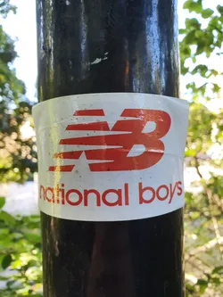 A sticker featuring the New Balance logo and the words 'national boys' affixed to a dark pole outdoors.