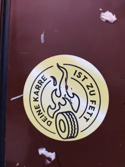 Round yellow sticker with a black outline. It shows a tire with flames and the text "Deine Karre ist zu fett" which translates from German to "Your car is too fat".