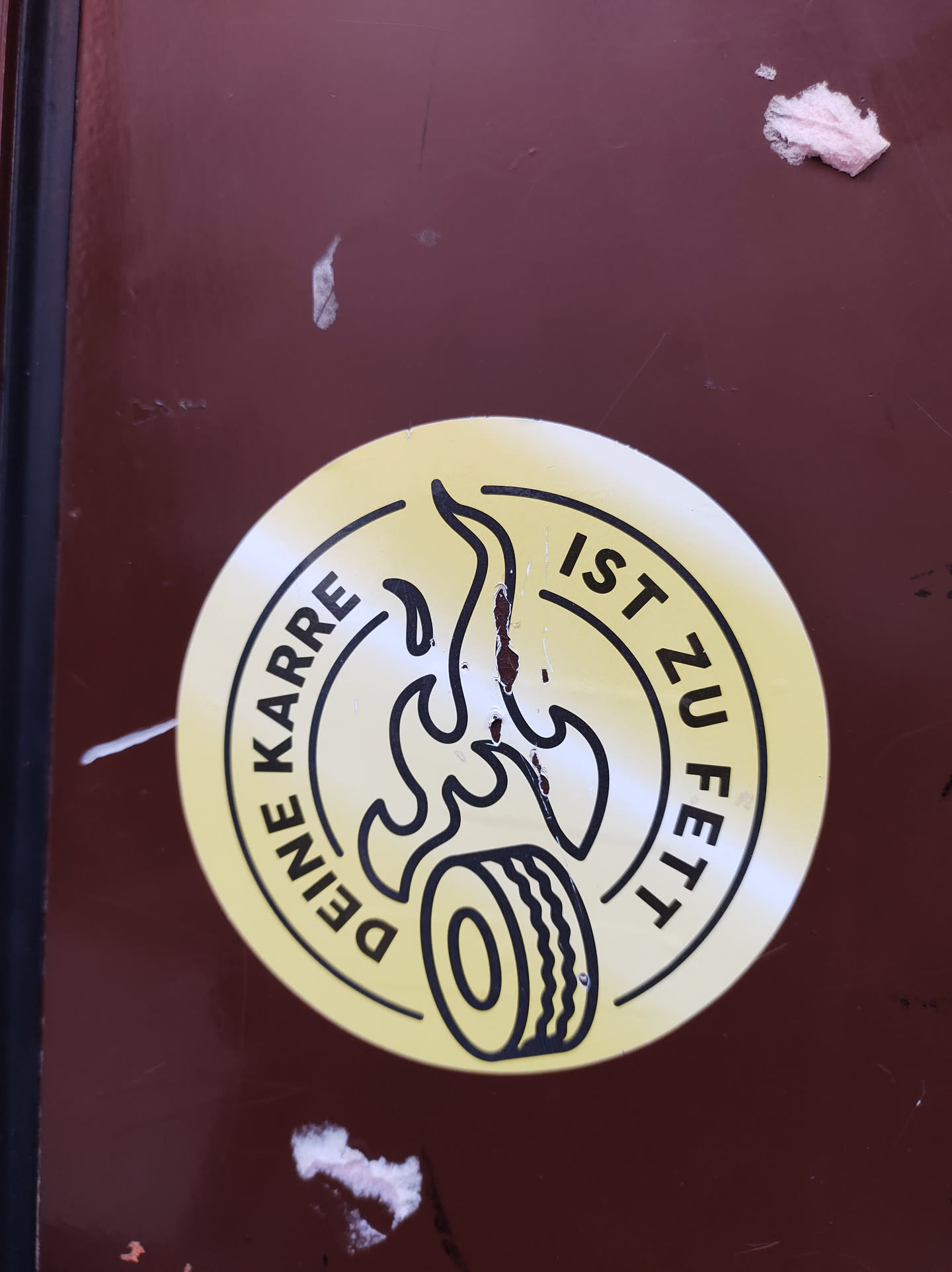 Round yellow sticker with a black outline. It shows a tire with flames and the text "Deine Karre ist zu fett" which translates from German to "Your car is too fat".