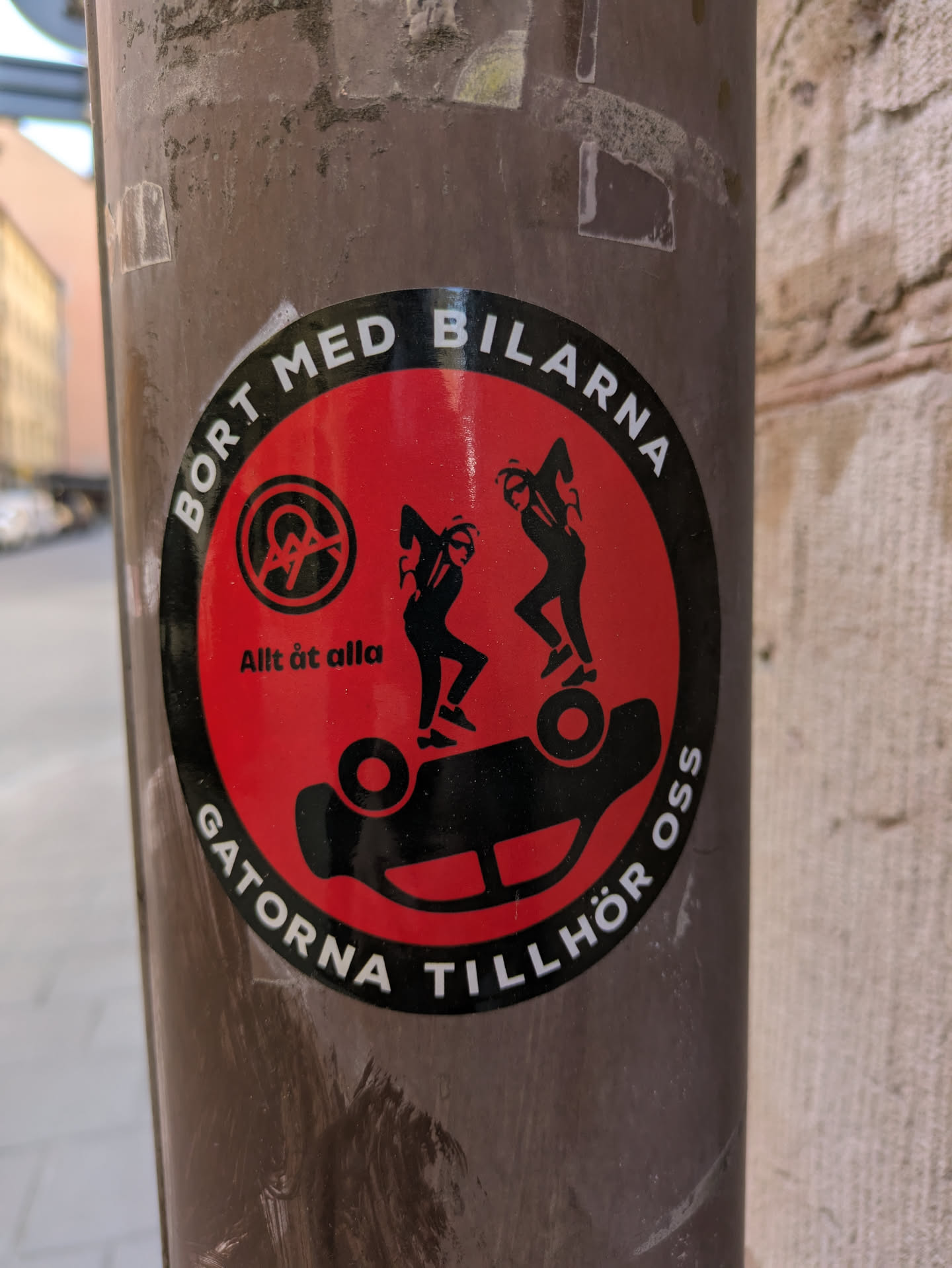 A round sticker on a pole. It features a red background with black silhouettes of two figures jumping on a car, which is flipped over. The text around the edge reads "BORT MED BILARNA" and "GATORNA TILLHÖR OSS" which translates to "Away with the cars" and "The streets belong to us". It also contains the text "Allt åt alla", and a symbol of crossed lines on a steering wheel.