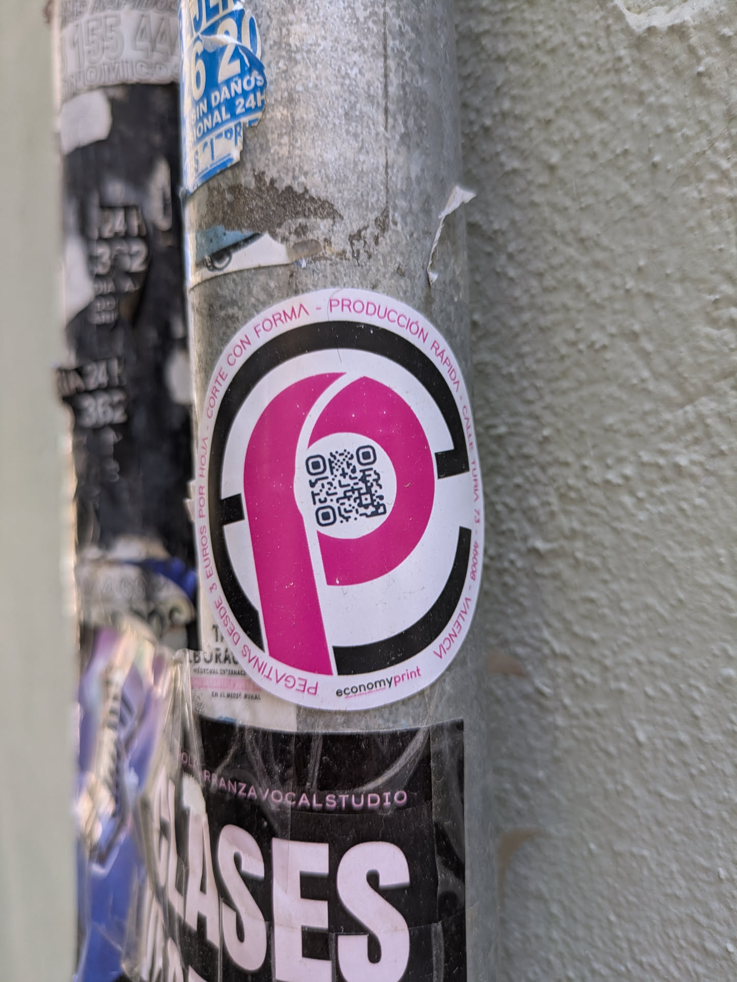 The sticker features a large pink 'P' inside a circle, with a QR code within the 'P'. The text around the circle includes "CORTE CON FORMA", "PRODUCCION RAPIDA", "CALLE TURIA 73", and "VALENCIA". The company name "economyprint" is printed at the bottom of the sticker. The sticker is attached to a weathered, gray pole.