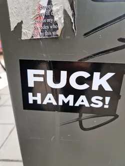 A black sticker with white bold text saying "FUCK HAMAS!" is affixed to a gray surface.  The sticker is rectangular and has a reflective surface. The background shows other stickers and graffiti.