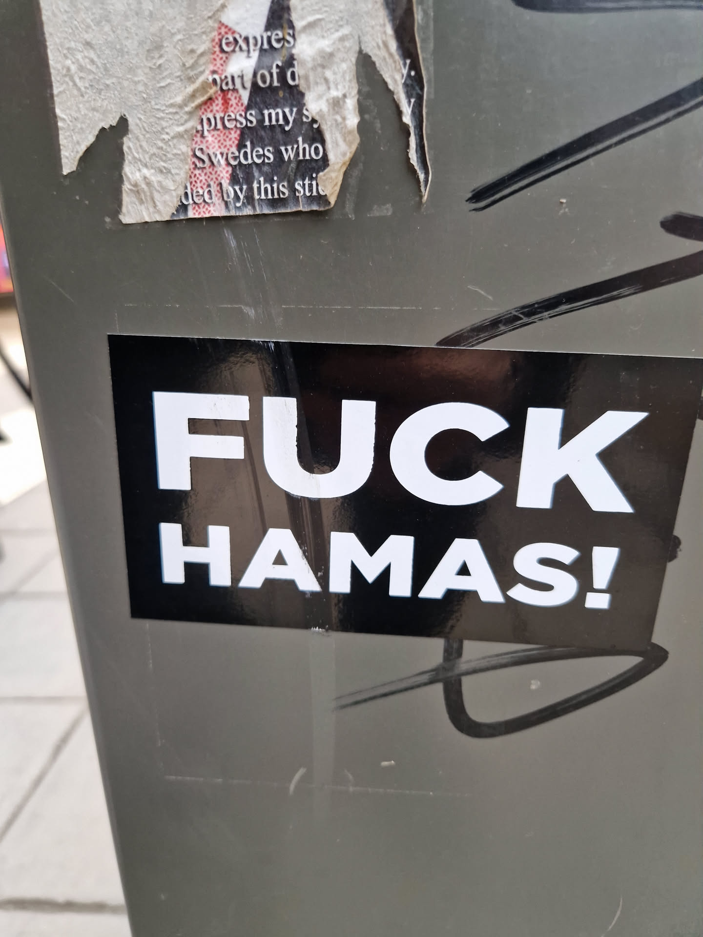 A black sticker with white bold text saying "FUCK HAMAS!" is affixed to a gray surface.  The sticker is rectangular and has a reflective surface. The background shows other stickers and graffiti.