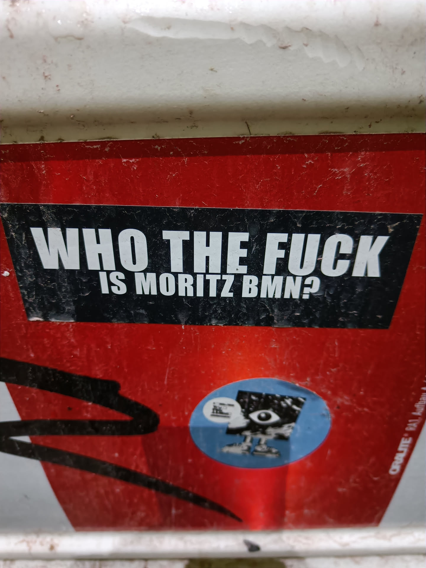A sticker with a provocative question. The sticker has a black background with white text and is pasted on a red surface. There is another smaller sticker next to it with a cartoon drawing of an eye.